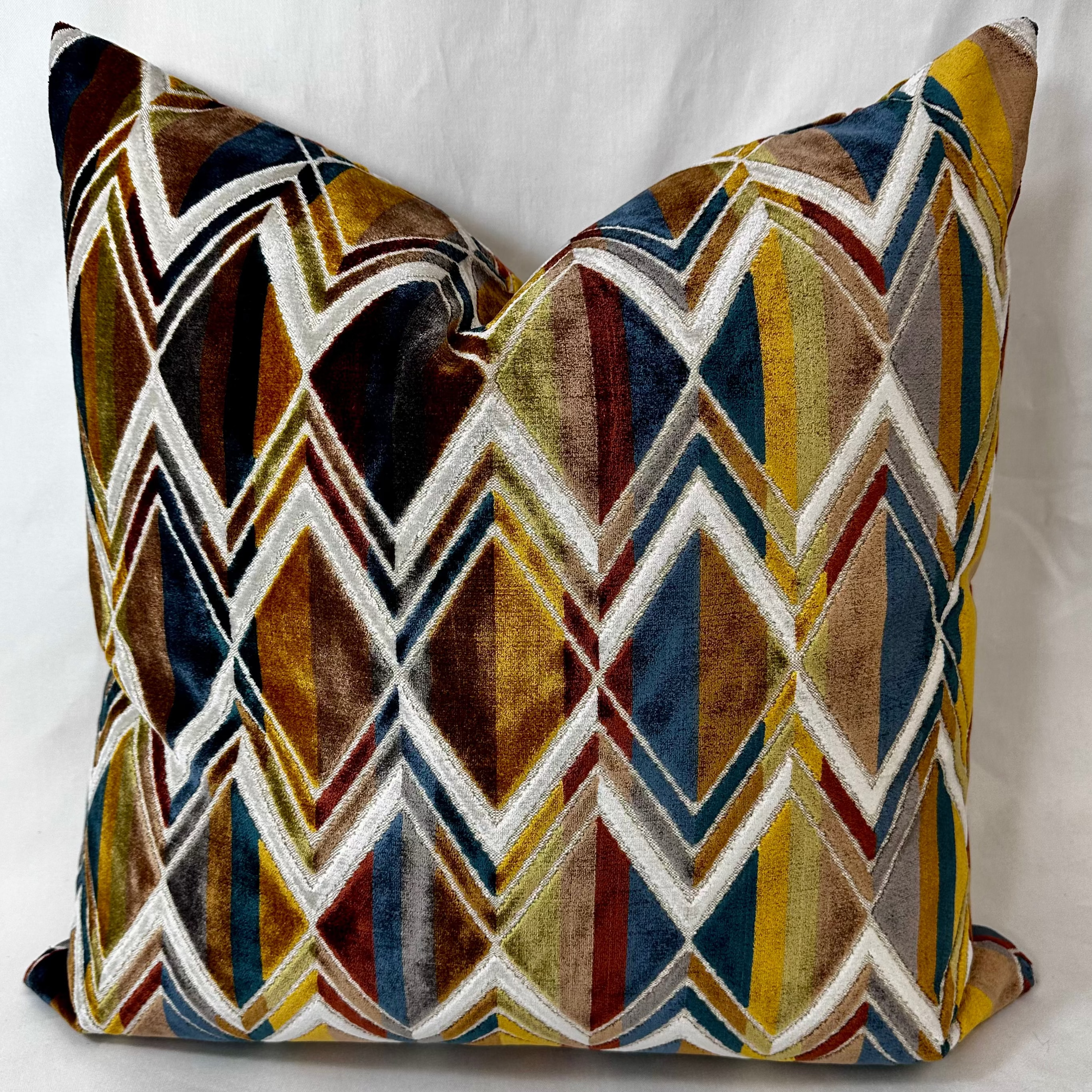 A square decorative throw pillow featuring a bold geometric chevron pattern in mustard yellow, navy blue, brown, burgundy, and white, set against a plain white fabric background.