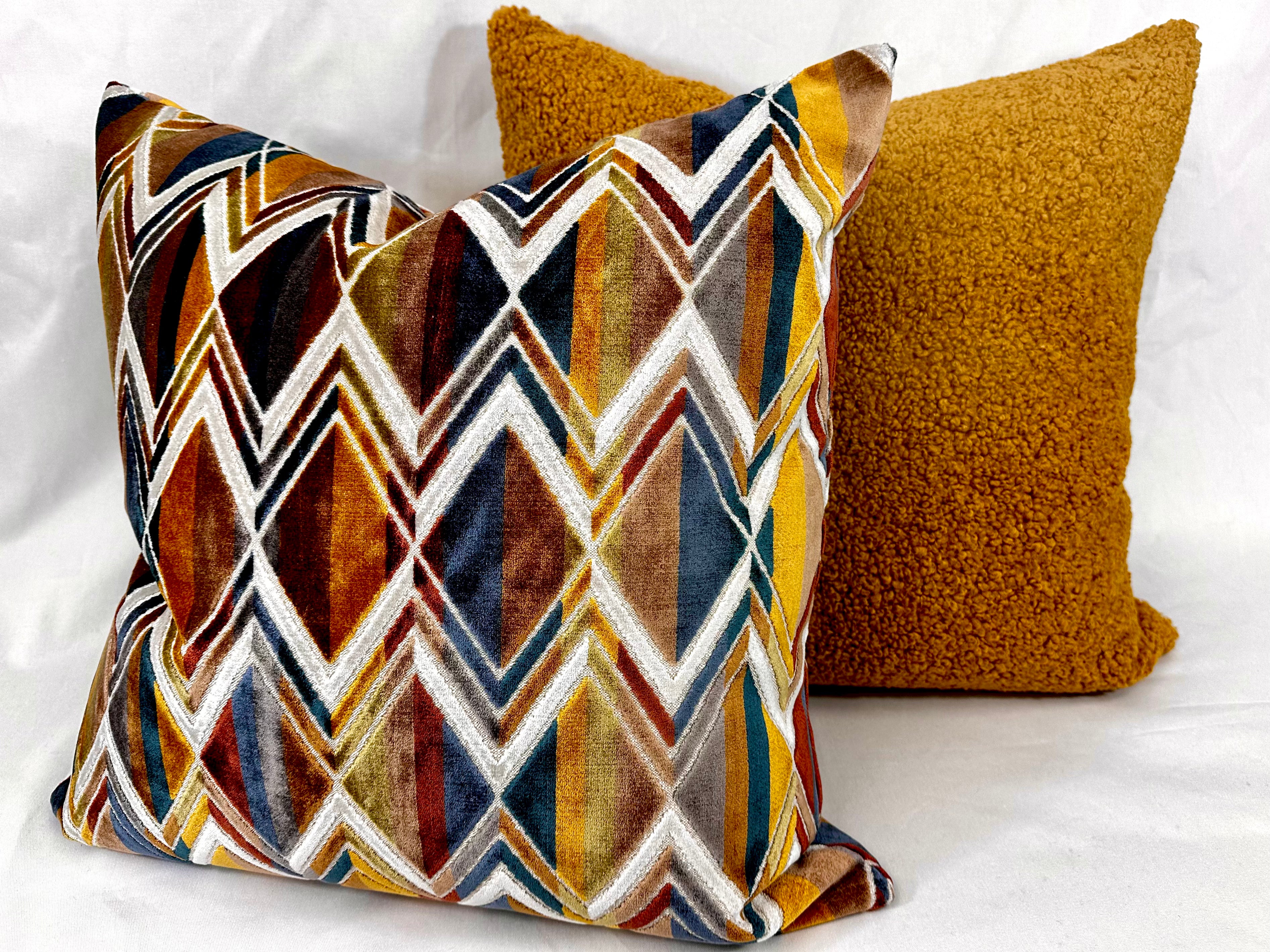 Two decorative throw pillows on a white fabric background. The front pillow features a multicolor geometric diamond pattern in brown, gold, navy, white, and red, while the back pillow is a solid textured mustard yellow.