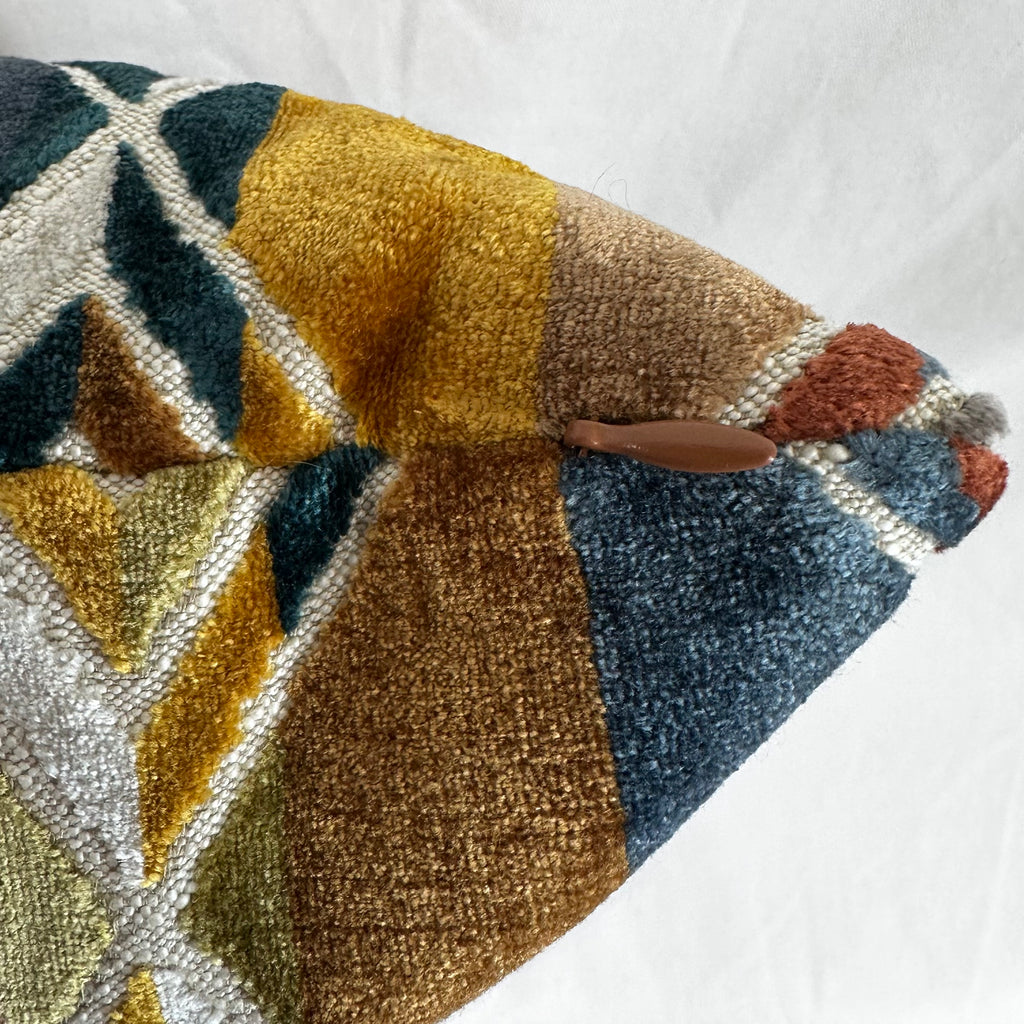 A close-up of a woven throw pillow cover featuring a geometric pattern in mustard yellow, brown, slate blue, cream, and rust, with a small brown leather accent tab.
