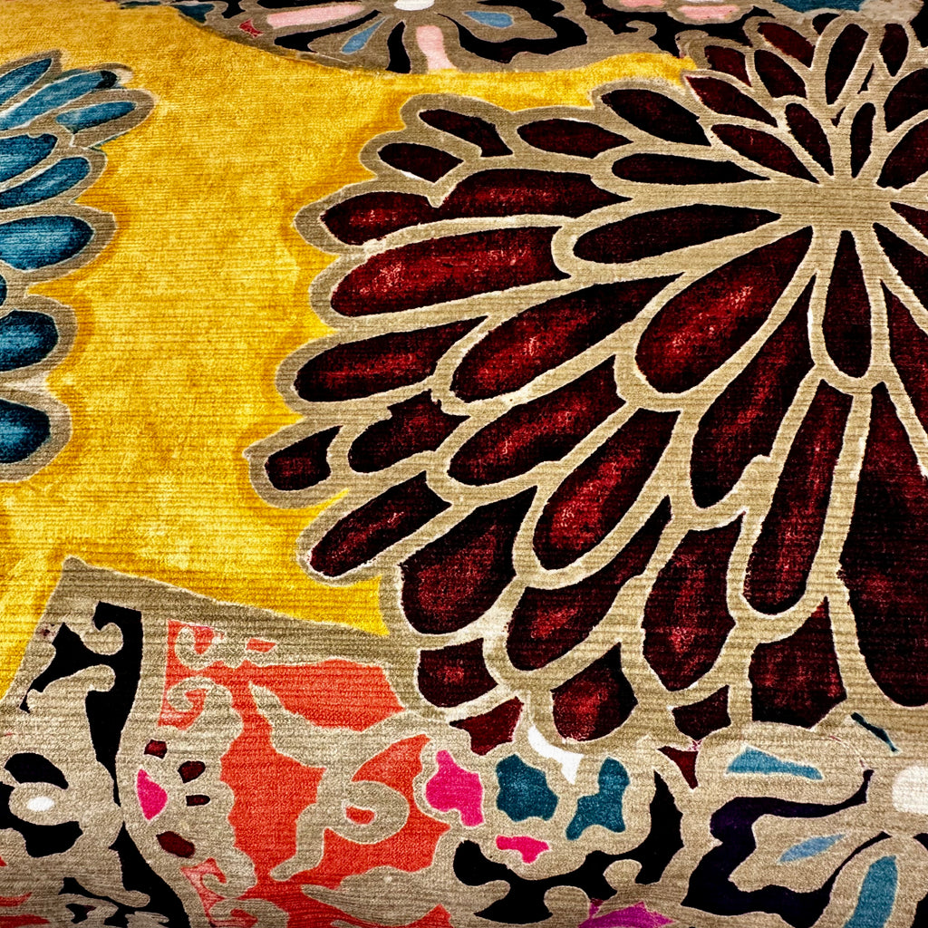 Close-up of a patterned textile or fabric with a large dark red floral motif outlined in beige on a golden yellow background, plus smaller multicolor abstract sections in pink, blue, black, and white.