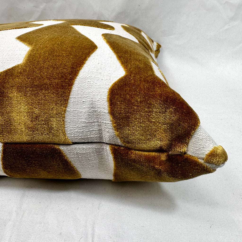 A close-up of a square throw pillow with a textured velvet-like finish, featuring a mustard yellow and white abstract pattern, resting on a white fabric background.