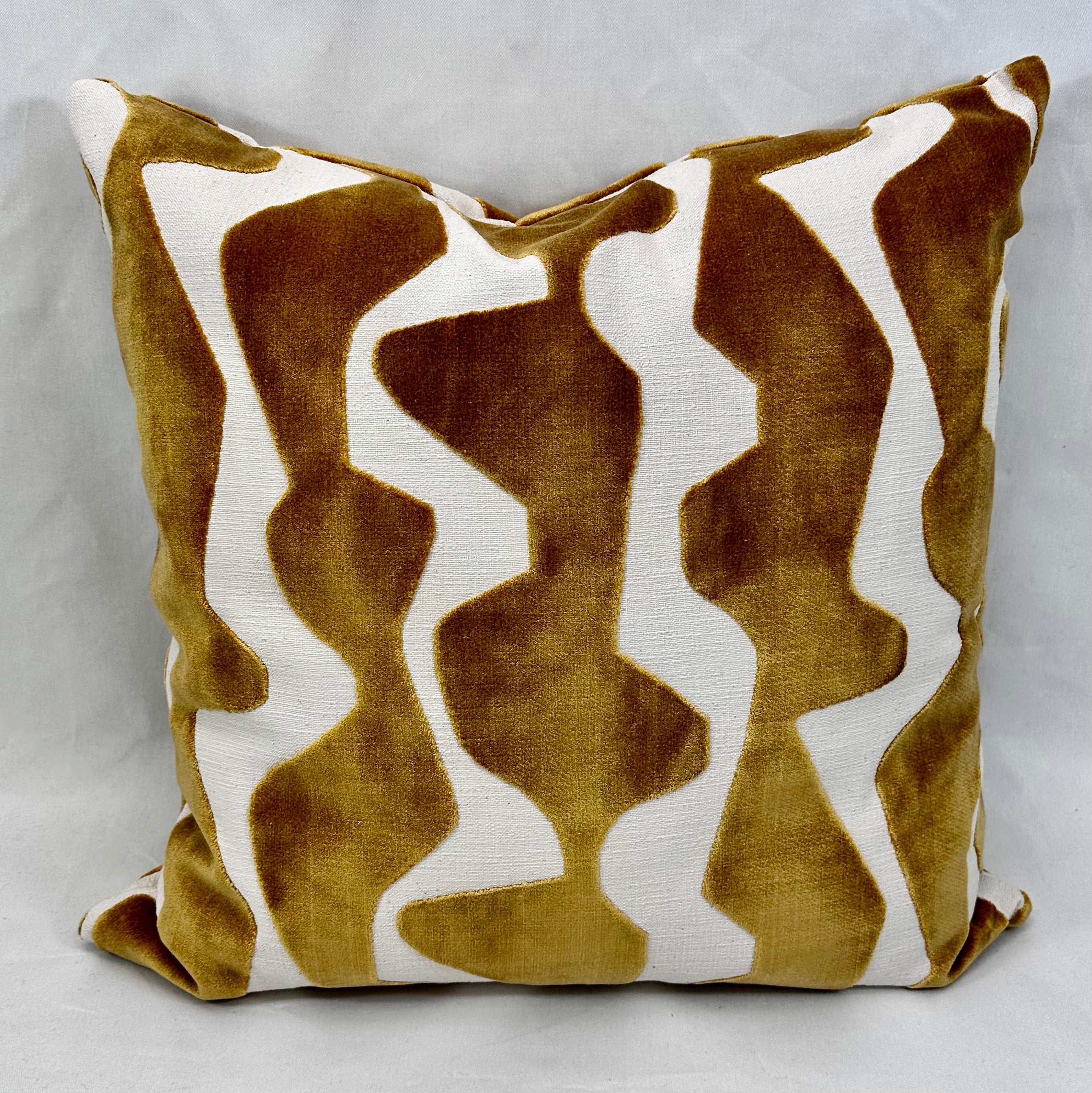 A square throw pillow featuring an abstract wavy pattern in mustard yellow velvet and off-white textured fabric, set against a neutral gray background.