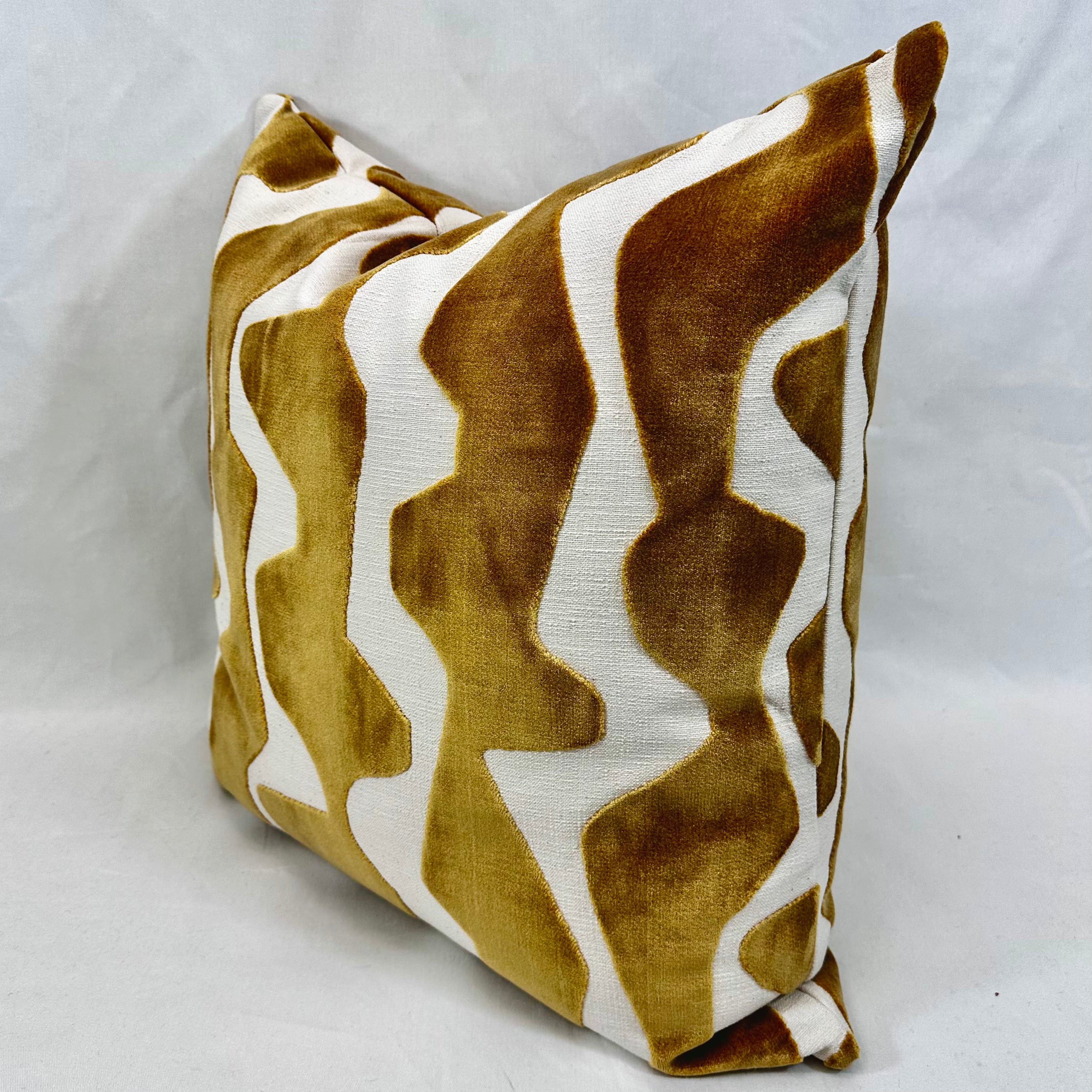 A square throw pillow featuring a bold, wavy abstract pattern in mustard yellow and white, photographed against a neutral light gray fabric backdrop.