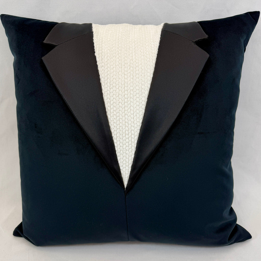 A square throw pillow designed to look like a tuxedo jacket, featuring a dark black velvet body with satin lapels and a textured cream knit center panel, set against a neutral light gray background.