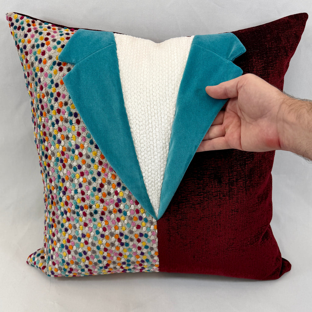 A square decorative throw pillow designed to look like a tuxedo jacket, featuring a multicolor polka-dot pattern on one side, a deep burgundy velvet side, and a white knit center with teal velvet lapels. A hand is holding the right lapel to show its texture and depth.