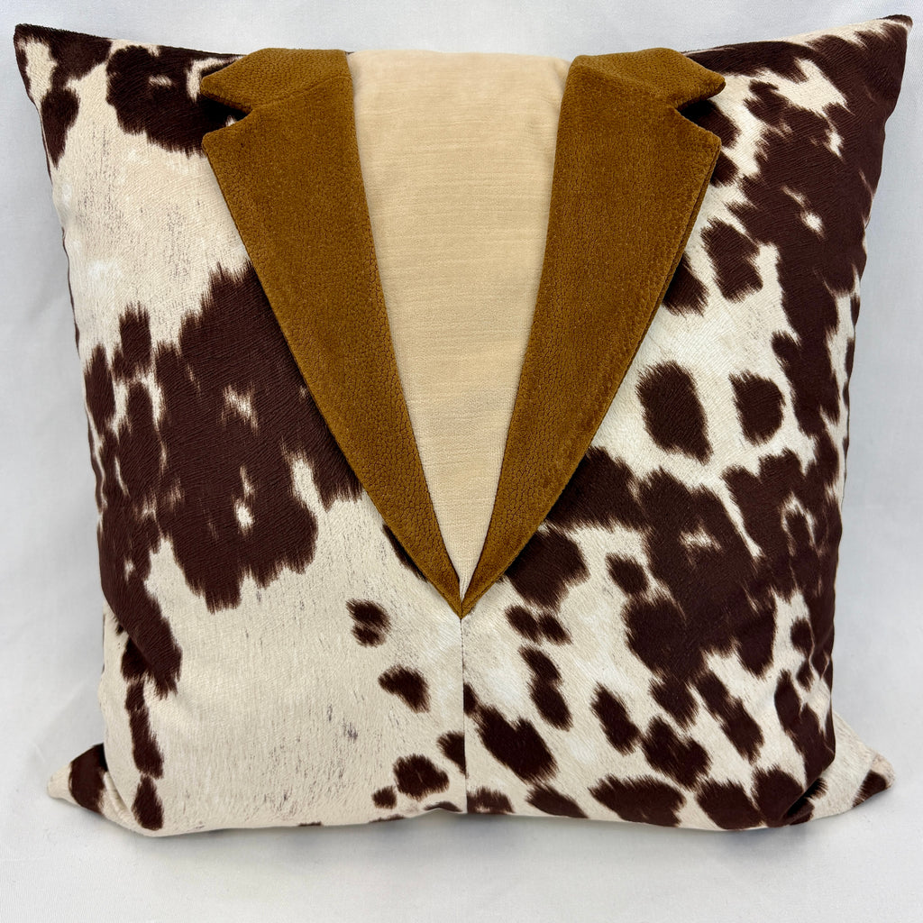 A square decorative throw pillow designed to look like a tuxedo jacket, featuring a brown and cream cowhide pattern on the body, a solid tan faux-suede lapel, and a cream fabric center panel.