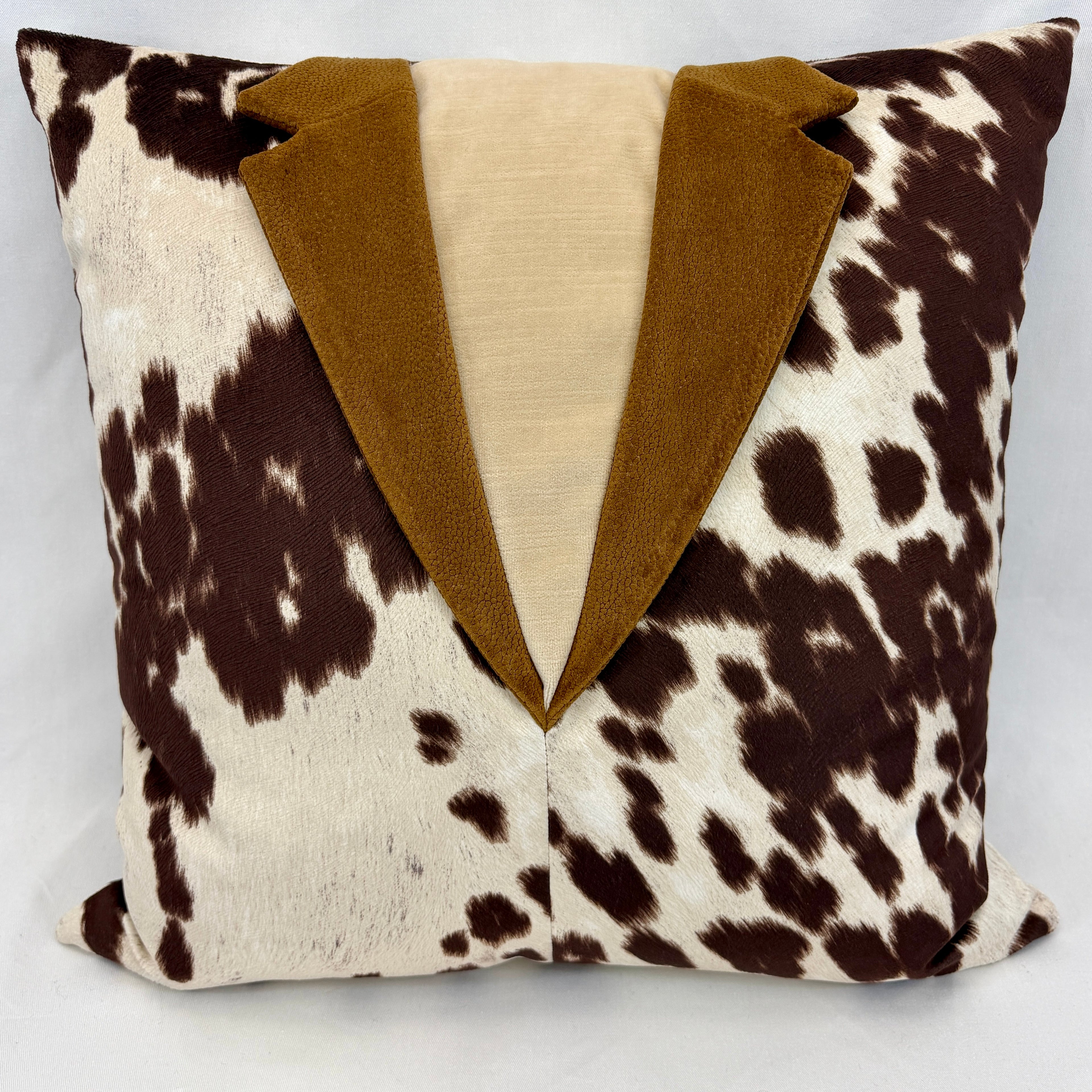 A square decorative throw pillow designed to look like a tuxedo jacket, featuring a brown and cream cowhide pattern on the body, a solid tan faux-suede lapel, and a cream fabric center panel.