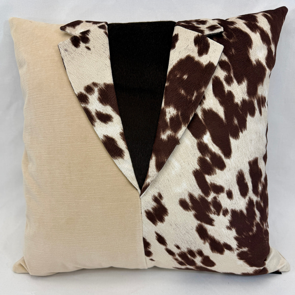 A square decorative throw pillow with a unique split design: one half is solid beige linen, and the other half features a brown and cream cow print. The center of the pillow is open, revealing a black plush lining behind the collar-like split.