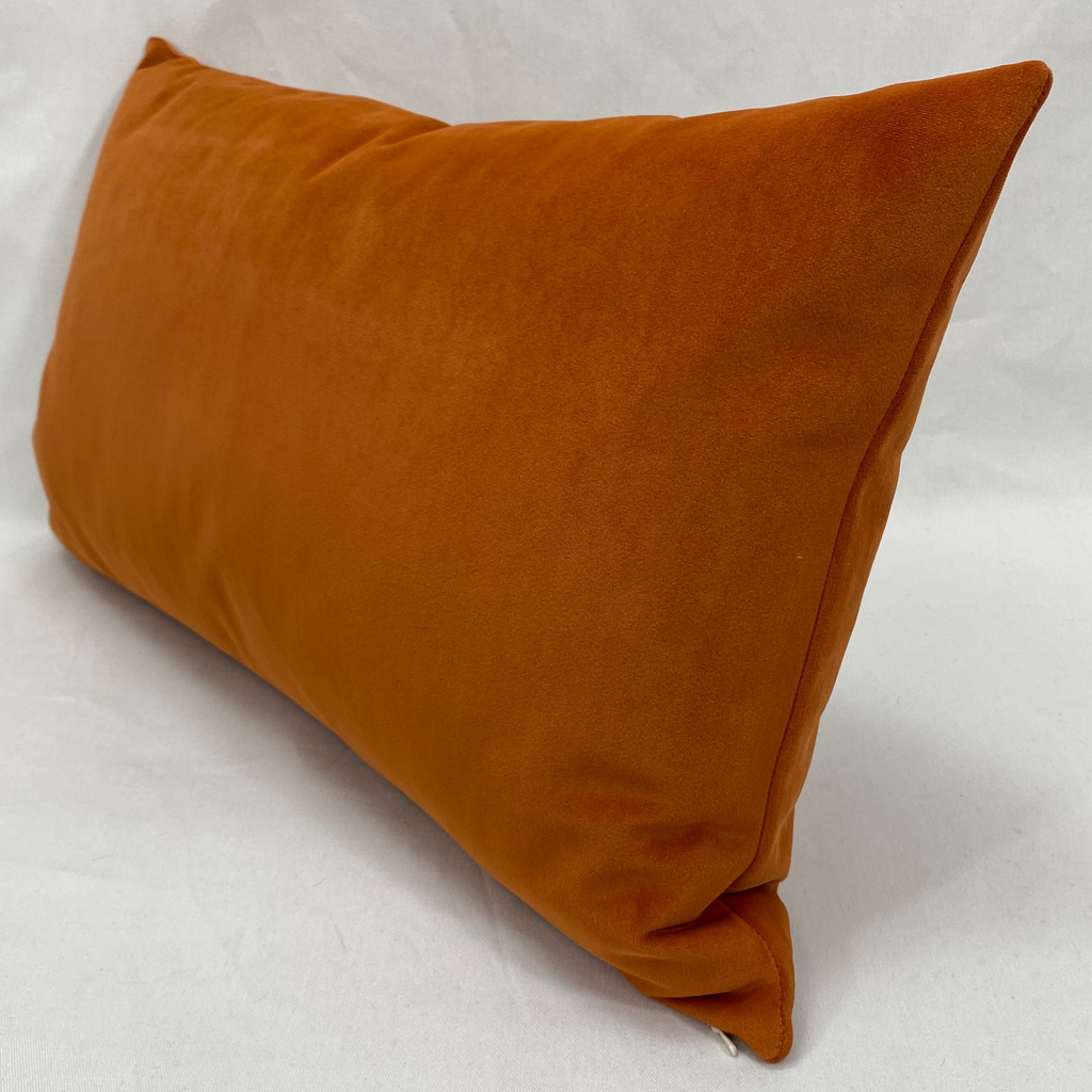 A rectangular lumbar throw pillow in a warm rust-orange velvet fabric, photographed at an angle against a white fabric backdrop.