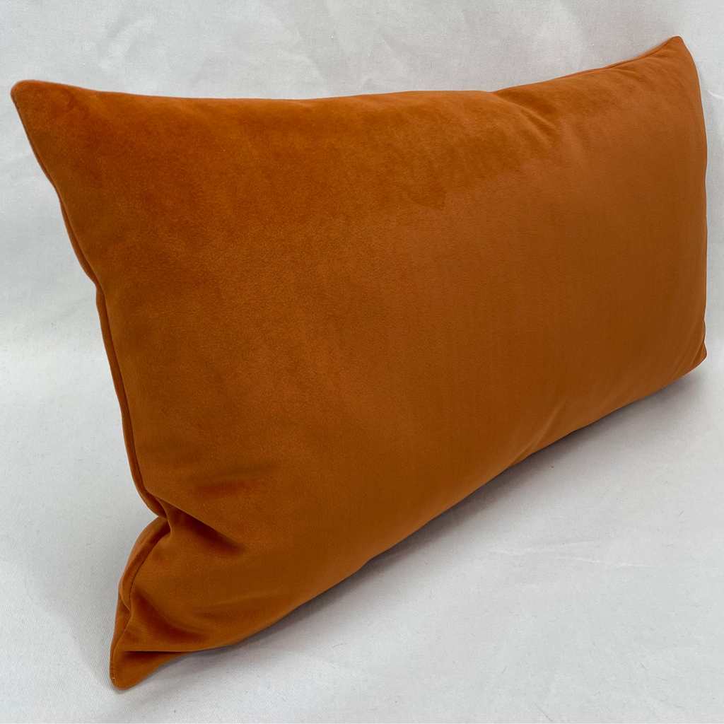A rectangular lumbar pillow in a warm rust-orange velvet fabric, photographed at an angle against a textured white backdrop.