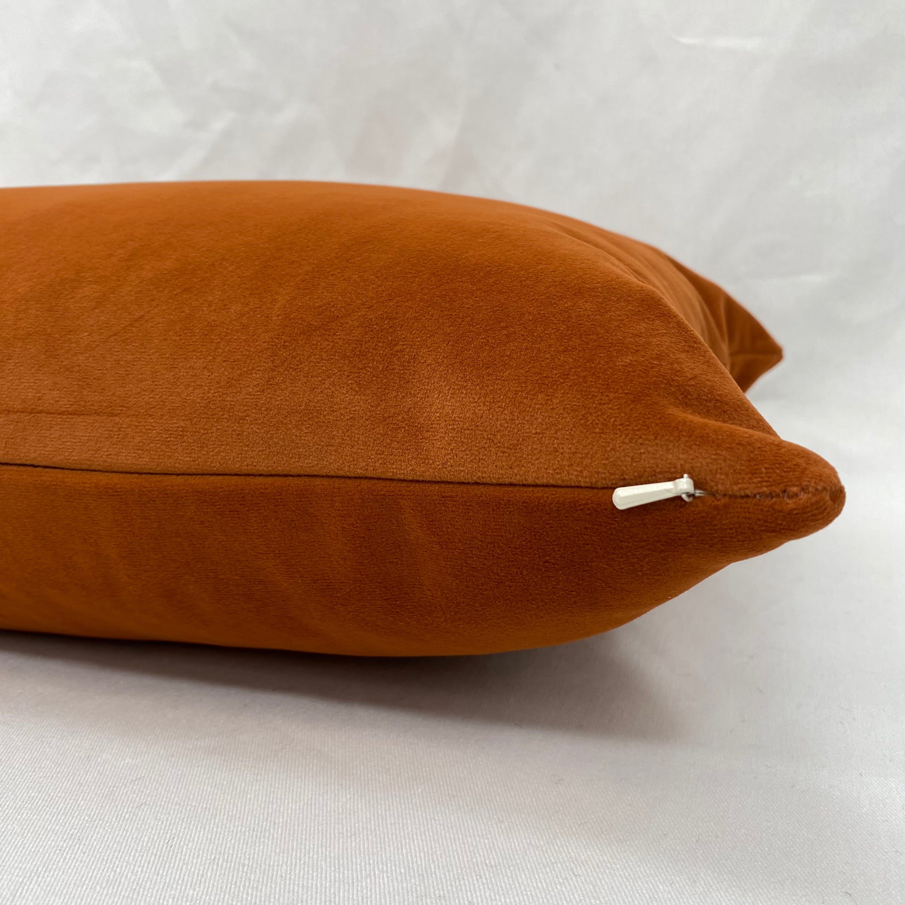 A close-up of a rectangular throw pillow in a warm rust-orange color, made from a soft suede-like fabric, with a white zipper pull visible on the right side.