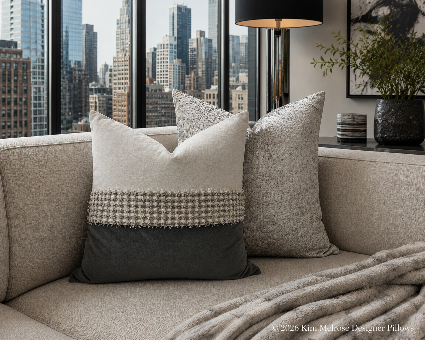 Two decorative throw pillows arranged on a light beige sofa in a modern living room setting with a city skyline visible through the windows. The front pillow is off-white on top, dark charcoal gray on the bottom, and features a textured beige and white houndstooth stripe across the middle.
