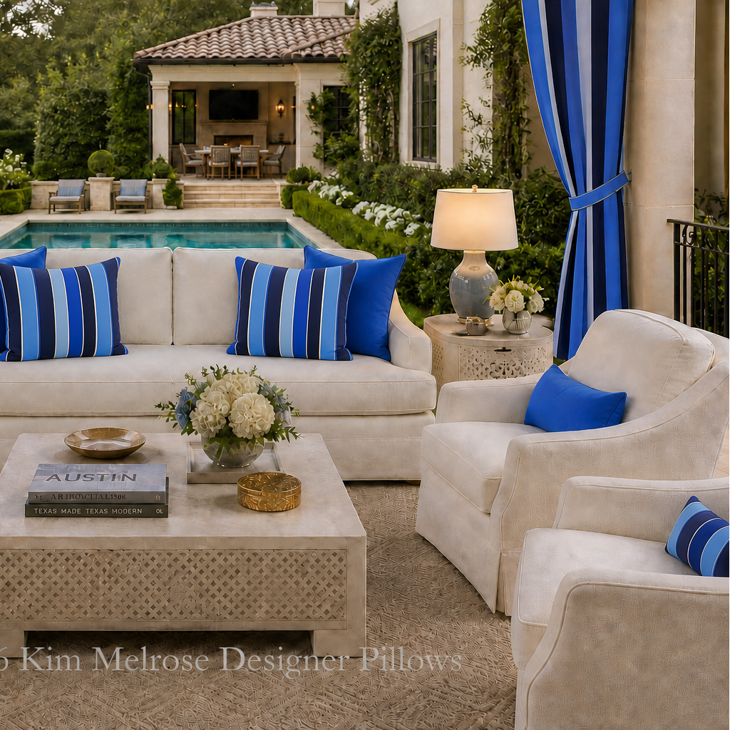 An outdoor patio seating area featuring a cream bouclé sofa and matching armchairs with blue and white striped pillows, arranged around a woven coffee table in front of a pool with a house and greenery in the background.