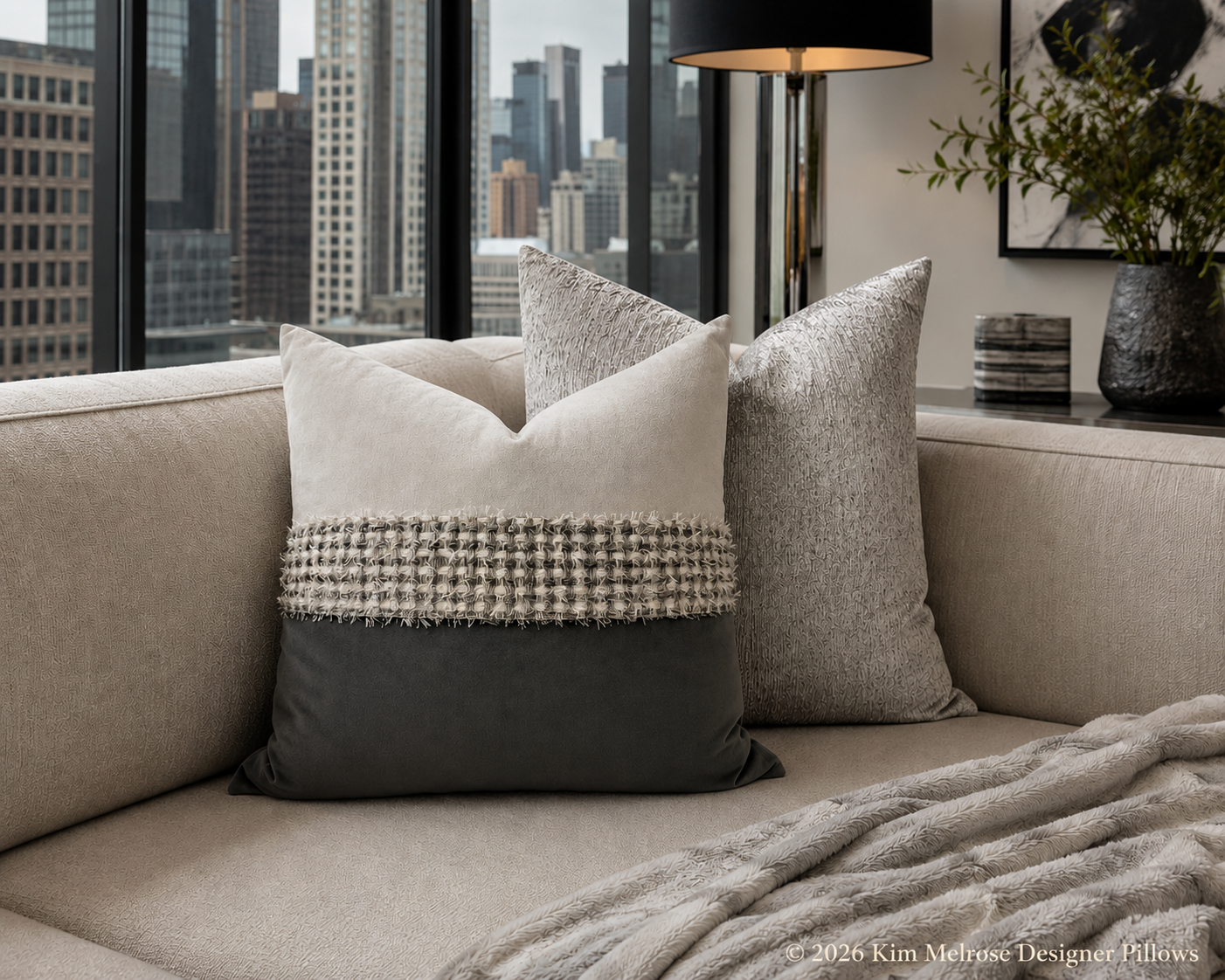 Two decorative throw pillows arranged on a beige sofa in a modern living room setting with a city skyline view. The front pillow is cream and dark gray with a textured woven band across the middle; the other pillow is heather gray.