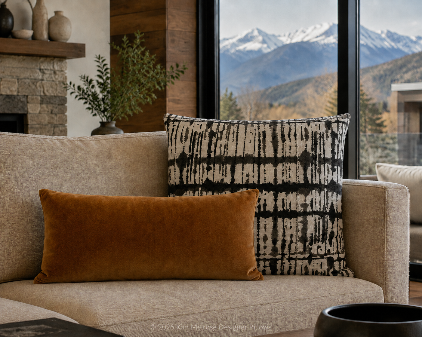 Two decorative throw pillows on a beige sofa in a modern living room setting with a large window showing a mountain landscape. The front pillow is a solid rust-orange velvet rectangle, and the back pillow features a black and off-white abstract vertical pattern.